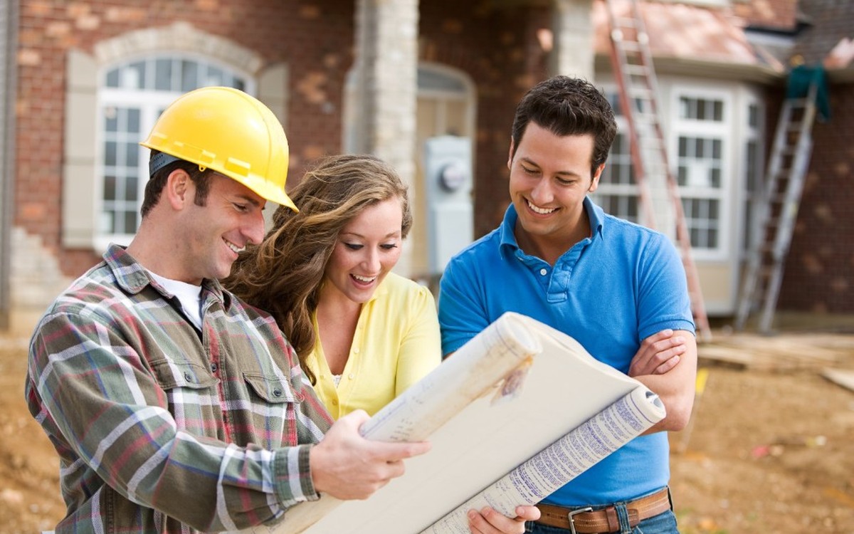 Construction professional reviewing blueprints with clients in front of a brick house being built.